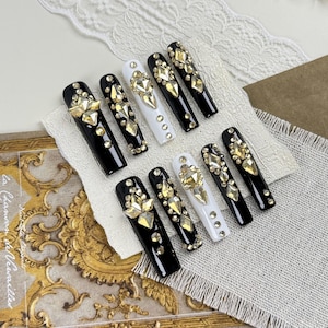 Op de afbeelding: Een set van tien lange, doodskistvormige valse nagels. De nagels zijn zwart en wit, versierd met goudkleurige strass-steentjes in verschillende maten en vormen. Sommige nagels hebben een grote, centrale edelsteen.