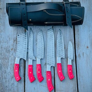 Puede incluir: Un juego de siete cuchillos de cocina de acero inoxidable con mangos de madera rojos, exhibidos con un estuche de cuero negro. Las hojas tienen una textura martillada. Los cuchillos est&aacute;n dispuestos sobre una superficie de madera envejecida.