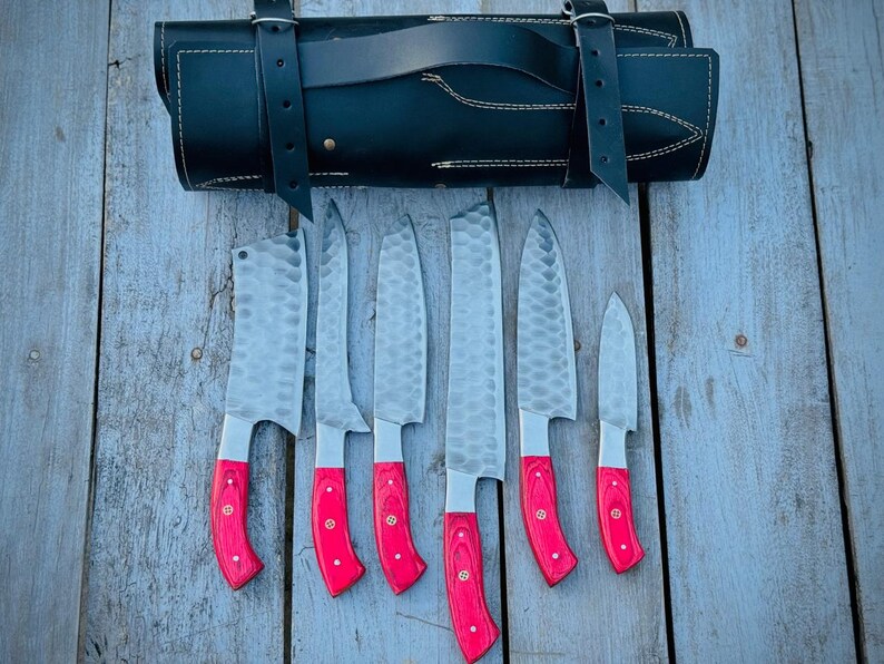 Puede incluir: Un juego de siete cuchillos de cocina de acero inoxidable con mangos de madera rojos, exhibidos con un estuche de cuero negro. Las hojas tienen una textura martillada. Los cuchillos est&aacute;n dispuestos sobre una superficie de madera envejecida.