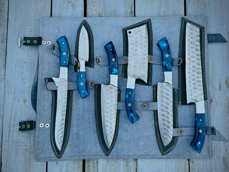 Puede incluir: Un juego de seis cuchillos de cocina con hojas de acero martillado y mangos de resina azul, presentados en un estuche de cuero gris. Los cuchillos incluyen una cuchilla y cuchillos de cocina. El estuche tiene un cierre de hebilla.