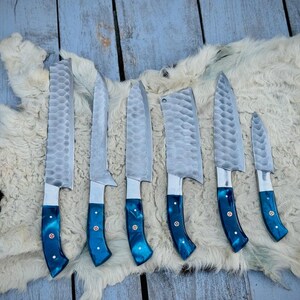 Puede incluir: Un juego de siete cuchillos de cocina con hojas plateadas texturizadas y mangos azules vibrantes. Los cuchillos est&aacute;n dispuestos sobre una piel blanca y marr&oacute;n, mostrando sus diferentes tama&ntilde;os y formas, adecuados para diversas tareas culinarias.