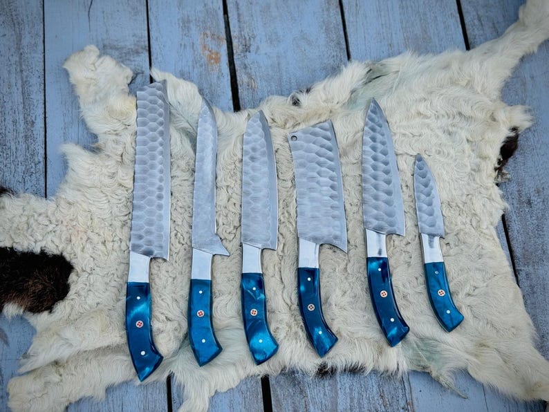 Puede incluir: Un juego de siete cuchillos de cocina con hojas plateadas texturizadas y mangos azules vibrantes. Los cuchillos est&aacute;n dispuestos sobre una piel blanca y marr&oacute;n, mostrando sus diferentes tama&ntilde;os y formas, adecuados para diversas tareas culinarias.