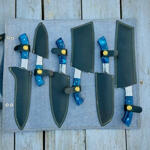 Puede incluir: Un juego de seis cuchillos con mangos azules y hojas plateadas, cada uno en una funda de cuero negro. Los cuchillos est&aacute;n dispuestos sobre un rollo de cuchillos gris, con correas y hebillas negras. Sobre una superficie de madera envejecida.