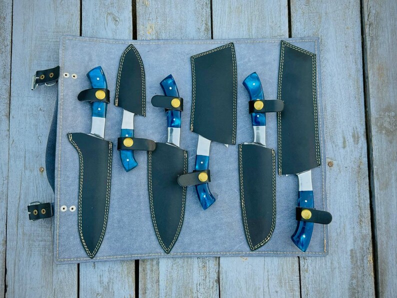 Puede incluir: Un juego de seis cuchillos con mangos azules y hojas plateadas, cada uno en una funda de cuero negro. Los cuchillos est&aacute;n dispuestos sobre un rollo de cuchillos gris, con correas y hebillas negras. Sobre una superficie de madera envejecida.