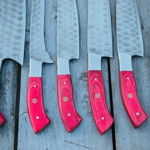 Puede incluir: Un conjunto de cuchillos de cocina con hojas de acero martillado y mangos de madera roja brillante. Los mangos est&aacute;n detallados con peque&ntilde;os remaches de lat&oacute;n. Los cuchillos se muestran sobre una superficie de madera envejecida.