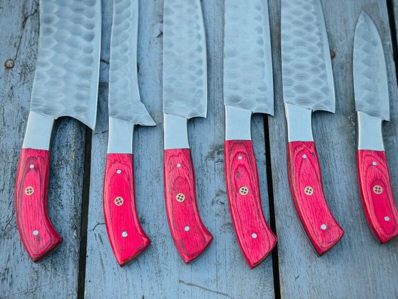 Puede incluir: Un conjunto de cuchillos de cocina con hojas de acero martillado y mangos de madera roja brillante. Los mangos est&aacute;n detallados con peque&ntilde;os remaches de lat&oacute;n. Los cuchillos se muestran sobre una superficie de madera envejecida.