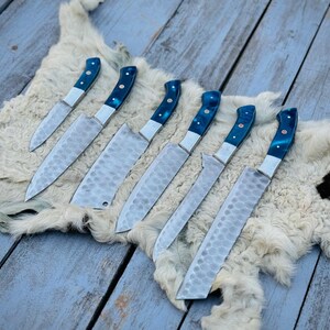 Puede incluir: Un conjunto de siete cuchillos de cocina con hojas texturizadas y mangos azules y plateados. Los cuchillos est&aacute;n colocados sobre una superficie similar a piel blanca, sobre un fondo de tablas de madera pintadas de azul desgastado. Los cuchillos var&iacute;an en tama&ntilde;o.