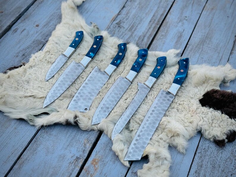 Puede incluir: Un conjunto de siete cuchillos de cocina con hojas texturizadas y mangos azules y plateados. Los cuchillos est&aacute;n colocados sobre una superficie similar a piel blanca, sobre un fondo de tablas de madera pintadas de azul desgastado. Los cuchillos var&iacute;an en tama&ntilde;o.
