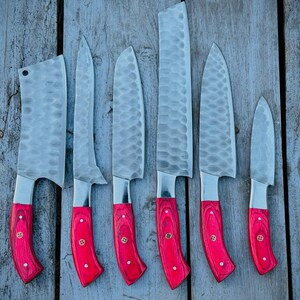 Puede incluir: Un juego de seis cuchillos de cocina con hojas de acero inoxidable y mangos de madera roja brillante. Las hojas tienen un acabado texturizado y los mangos est&aacute;n asegurados con remaches de lat&oacute;n. Los cuchillos est&aacute;n colocados sobre una superficie de madera envejecida.