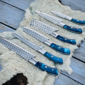 Puede incluir: Un juego de seis cuchillos de cocina con hojas plateadas texturizadas y mangos azules vibrantes. Los cuchillos est&aacute;n colocados sobre una superficie blanca similar a la piel, sobre un fondo de madera azul claro envejecido. Los mangos tienen un efecto veteado.