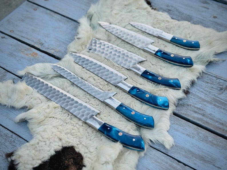 Puede incluir: Un juego de seis cuchillos de cocina con hojas plateadas texturizadas y mangos azules vibrantes. Los cuchillos est&aacute;n colocados sobre una superficie blanca similar a la piel, sobre un fondo de madera azul claro envejecido. Los mangos tienen un efecto veteado.