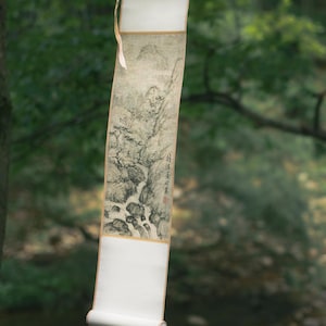 Peut inclure: Un rouleau suspendu avec une peinture de paysage en noir et blanc, représentant une cascade et des montagnes. Le rouleau est suspendu à une branche d'arbre, avec un bord blanc et un bas enroulé. L'arrière-plan montre une forêt floue et un ruisseau.