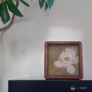 May include: A square, framed artwork featuring a delicate, light pink and white flower with green accents. The artwork is set against a tan background and enclosed in a dark wood frame. The bottom right corner of the frame has the text "X 吟翠堂".