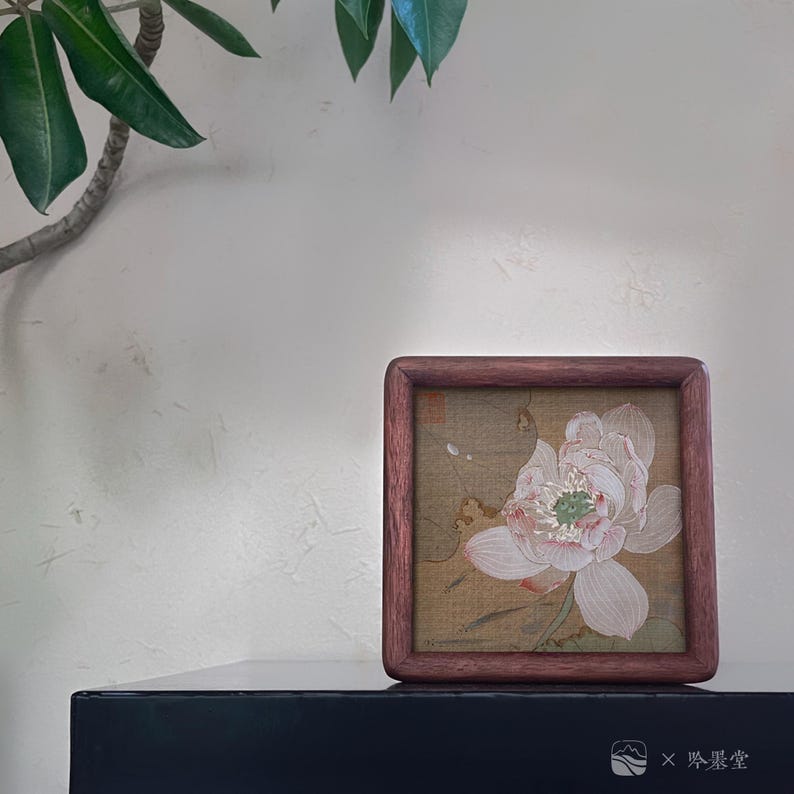 May include: A square, framed artwork featuring a delicate, light pink and white flower with green accents. The artwork is set against a tan background and enclosed in a dark wood frame. The bottom right corner of the frame has the text "X 吟翠堂".