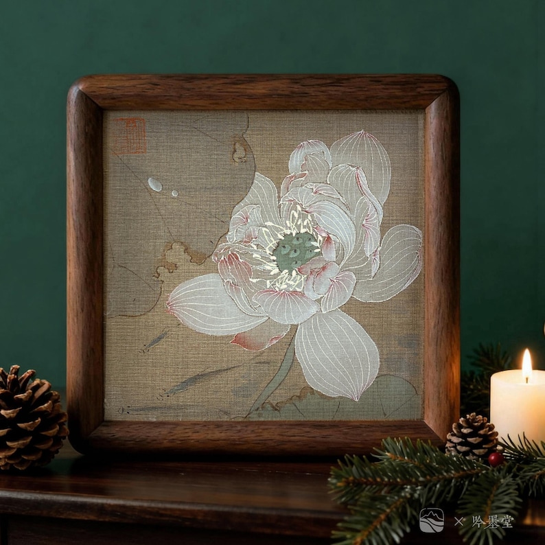 May include: A framed artwork featuring a detailed illustration of a white lotus flower with pink accents. The artwork is set against a beige background and framed in a dark brown wooden frame. The image is displayed on a dark wooden surface.