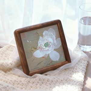 May include: Square wooden framed artwork featuring a detailed white lotus flower with pink and green accents. The artwork is set against a muted green background and rests on a cream-colored knitted surface. A glass of water is in the background.