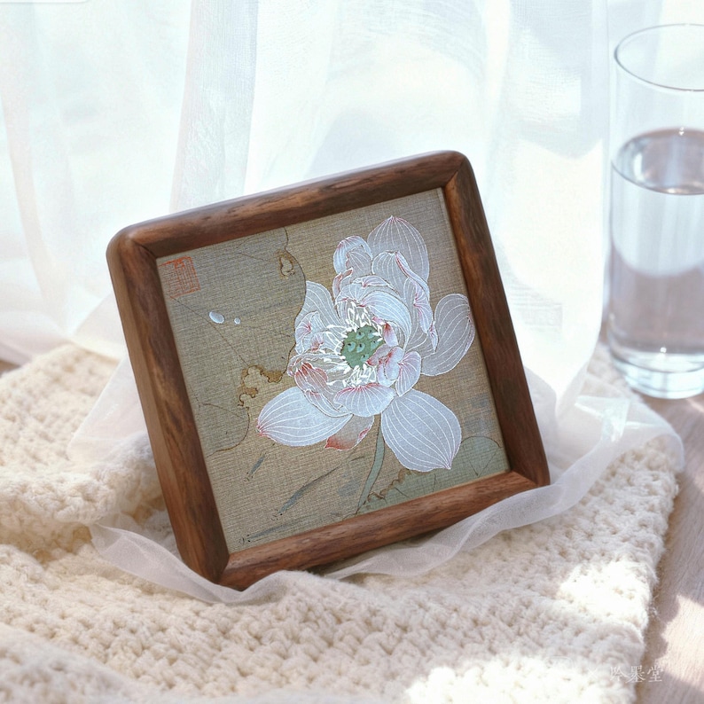 May include: Square wooden framed artwork featuring a detailed white lotus flower with pink and green accents. The artwork is set against a muted green background and rests on a cream-colored knitted surface. A glass of water is in the background.