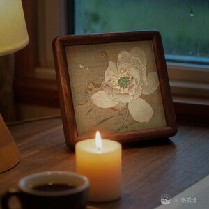 May include: A framed floral print featuring a lotus flower in soft pink and white hues, set against a muted background. The frame is made of dark wood. A lit candle and a cup of coffee sit on a wooden surface, creating a cozy atmosphere.