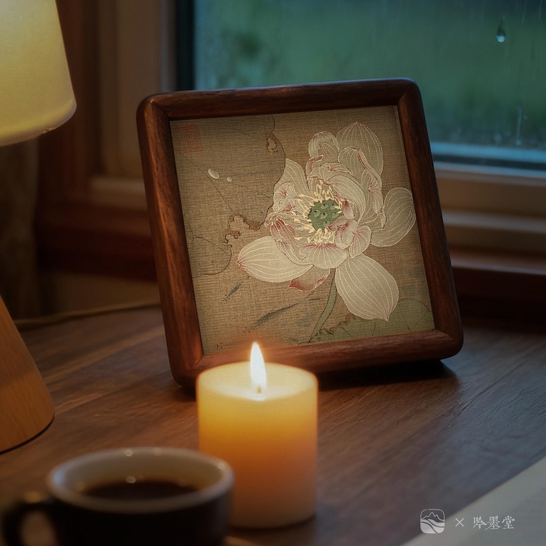 May include: A framed floral print featuring a lotus flower in soft pink and white hues, set against a muted background. The frame is made of dark wood. A lit candle and a cup of coffee sit on a wooden surface, creating a cozy atmosphere.