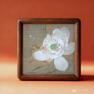 May include: Square wooden framed artwork featuring a detailed illustration of a white lotus flower with green center. The artwork is set against a beige background and framed in a dark brown wood. The image has a traditional Asian art style.