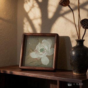 May include: A square, wooden-framed artwork featuring a delicate, white and green lotus flower illustration. The artwork rests on a dark wooden surface, next to a dark vase with dried stems and seed pods. The scene is bathed in warm light.