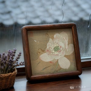 May include: A square wooden frame with rounded corners holds a floral print of a white lotus flower with pink and green accents. The frame is a dark brown color. A small basket of lavender sits to the left of the frame.