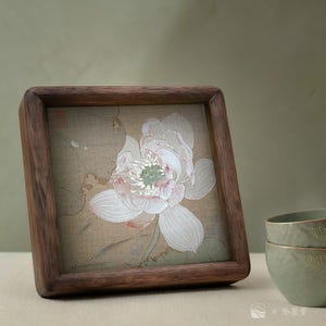 May include: A square wooden frame with a detailed illustration of a white lotus flower with pink accents. The artwork is set against a beige background. The frame is made of dark brown wood.