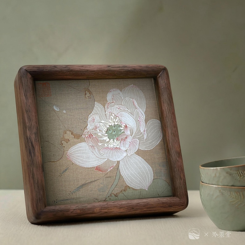 May include: A square wooden frame with a detailed illustration of a white lotus flower with pink accents. The artwork is set against a beige background. The frame is made of dark brown wood.