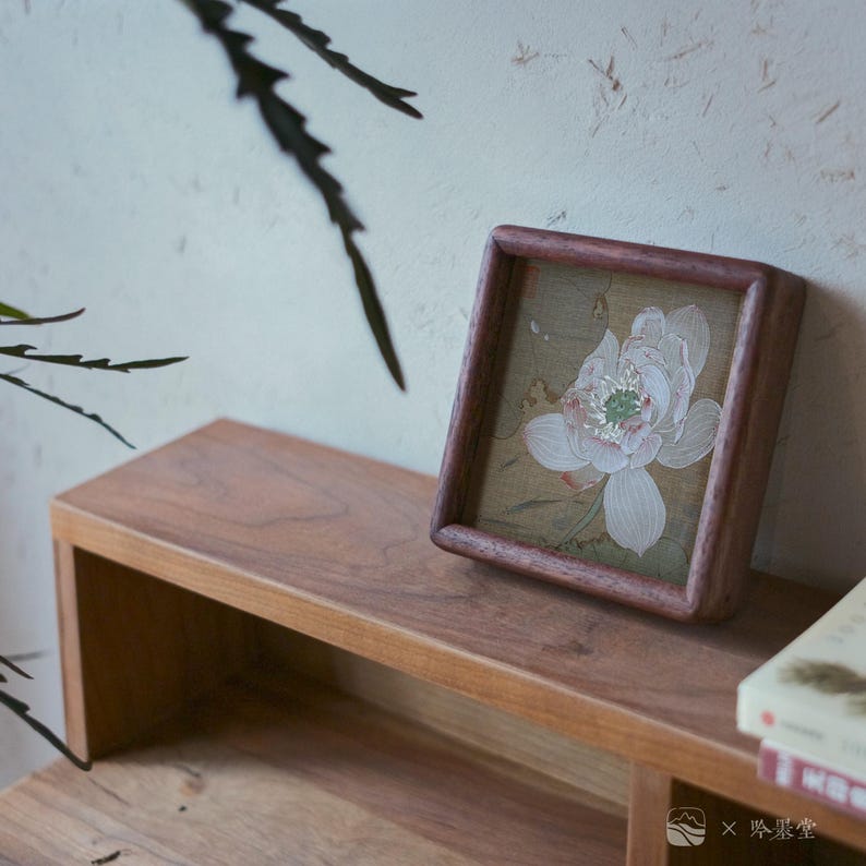 May include: A square, wooden framed artwork featuring a delicate white and pink lotus flower painting. The artwork is displayed on a wooden shelf, with a book visible in the background. The overall aesthetic is minimalist and serene.