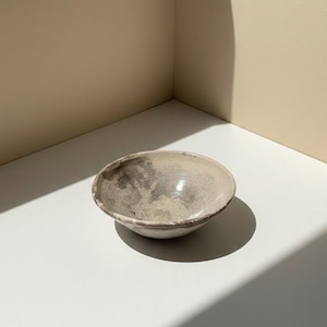 Pode incluir: Uma pequena tigela de cerâmica branca suja com uma forma ligeiramente irregular. A tigela tem uma superfície texturizada com marcas cinzentas e castanhas subtis. O interior é liso e a borda é ligeiramente mais escura. A tigela está numa superfície branca.