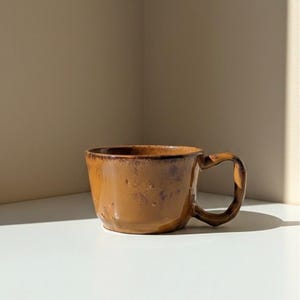 Pode incluir: Caneca de cerâmica castanha artesanal, com uma superfície texturizada única. A caneca apresenta um corpo arredondado e uma pega distinta. A cor é um tom quente e terroso, perfeito para desfrutar de café ou chá. O fundo é neutro.