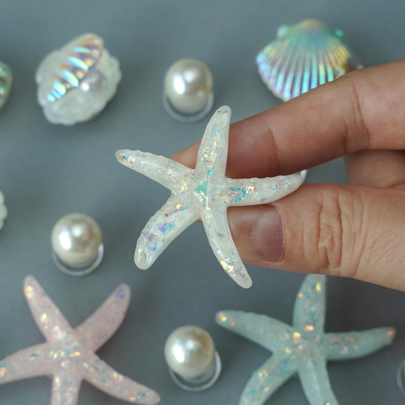 May include: A collection of iridescent starfish, seashells, and pearl beads. The starfish are white with shimmering glitter accents. The seashells have a pearlescent sheen, and the pearls are round and smooth.
