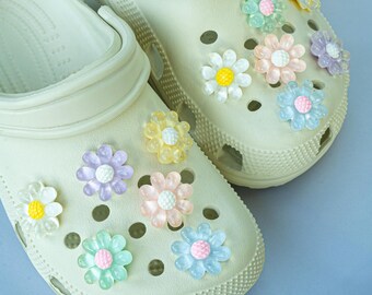May include: A pair of beige clogs adorned with colorful flower charms. The charms are in various pastel colors, including pink, blue, yellow, and purple, and are attached to the top of the clogs. The clogs are made of a rubbery material and have a textured surface.
