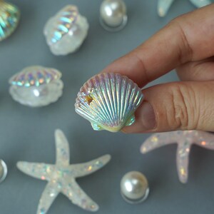 May include: Iridescent seashell with a small gold star, held by a hand. Other decorative elements include white starfish, pearls, and other seashells. The items have a pearlescent sheen, suggesting a coastal or nautical theme.