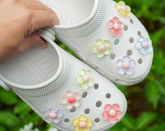 May include: A pair of white clogs decorated with colorful flower charms. The charms are in shades of pink, yellow, green, and blue. The clogs are made of a rubbery material and have a textured surface.