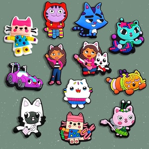 May include: A collection of colorful, cartoon-style cat and character charms. The charms feature various cat designs in bright colors, including pink, blue, and orange, and characters with brown hair. Some charms are in the shape of a car.