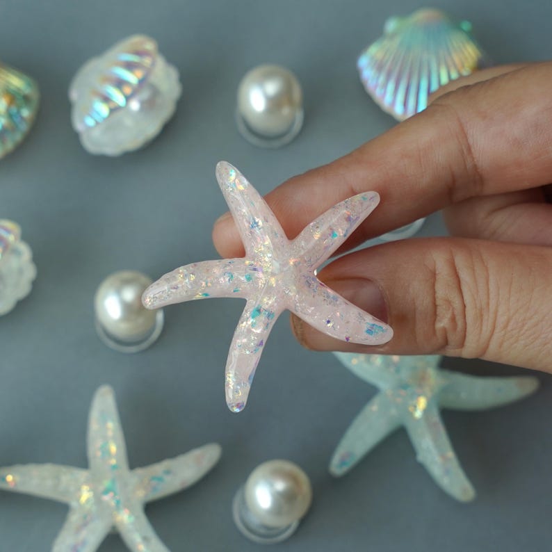 May include: A light pink starfish with iridescent glitter is held by a hand. Other starfish, seashells, and pearls are scattered on a gray surface. The starfish has five points and is slightly translucent.