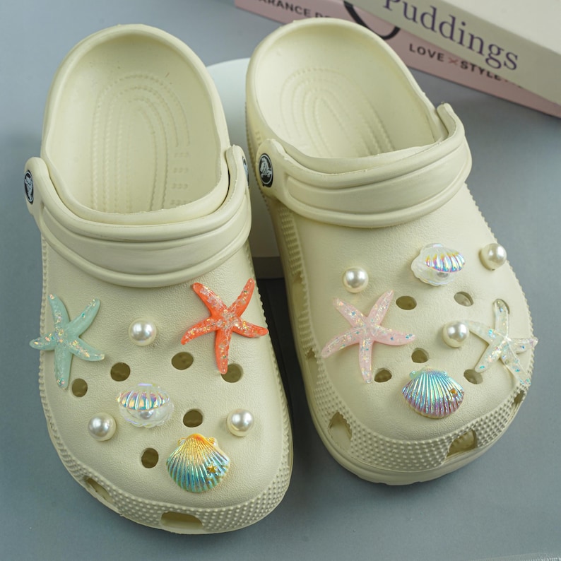 May include: A pair of cream-colored clogs decorated with starfish, seashells, and faux pearls. The starfish are in shades of orange, pink, and turquoise. The seashells have iridescent finishes.