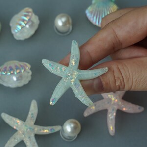 May include: A collection of iridescent seashell and starfish decorations. The starfish are light blue with glitter accents. Several white pearl beads are scattered around the starfish and seashells.