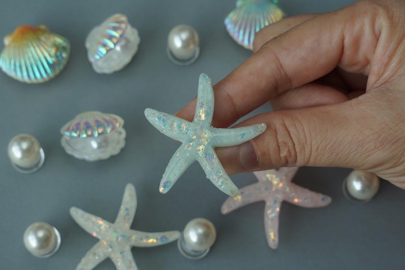 May include: A collection of iridescent seashell and starfish decorations. The starfish are light blue with glitter accents. Several white pearl beads are scattered around the starfish and seashells.
