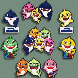 May include: A collection of colorful cartoon shark-shaped magnets. The magnets feature various colors, including blue, pink, yellow, green, and orange. Each shark has a smiling face and is holding a sign. The magnets are likely for decorative purposes.