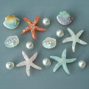May include: An assortment of decorative seashells, starfish, and faux pearls. The seashells are in shades of blue, green, and iridescent white. The starfish are orange, pink, and blue with glitter. The pearls are white.