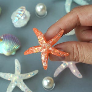 May include: An orange starfish with glitter accents is held by a hand. Other decorative elements include seashells, pearls, and other starfish. The starfish is a vibrant orange color with sparkling glitter embedded within.