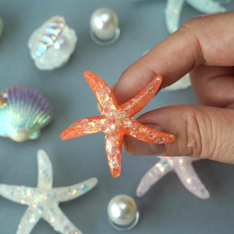 May include: An orange starfish with glitter accents is held by a hand. Other decorative elements include seashells, pearls, and other starfish. The starfish is a vibrant orange color with sparkling glitter embedded within.