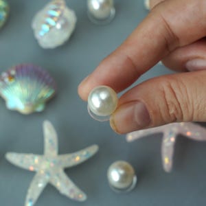 May include: A close-up of a pearl bead held by a hand, with other decorative seashells and starfish. The pearl is white and round, with a clear plastic backing. The other decorations are iridescent and glittery.