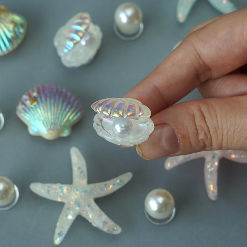 May include: Iridescent seashell and pearl embellishments. The image features several decorative elements, including open and closed seashells, pearls, and starfish. The seashells and starfish have a shimmering, glittery finish, and the pearls are round and white.