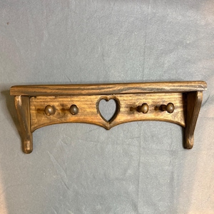 May include: Wooden wall shelf with a distressed finish. The shelf features a heart-shaped cutout and four wooden pegs for hanging items. The wood has a warm, brown tone with darker accents, creating a rustic aesthetic. The shelf is designed for wall mounting.