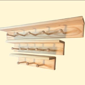 May include: Three wooden coat racks of varying sizes, each with a shelf and a row of wooden pegs. The racks are made of light-colored wood and are arranged in a stacked configuration, showcasing their different dimensions and design.