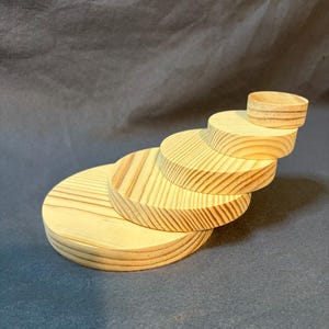 May include: A set of five unfinished, light-colored wooden circles stacked in a cascading arrangement. The wood grain is visible on each disc, with the smallest circle at the top. The arrangement is on a dark gray background.