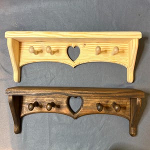May include: Two wooden wall shelves with a heart-shaped cutout and four pegs each. The top shelf is natural wood, while the bottom shelf is stained a dark brown. Both shelves have a decorative, scalloped edge.